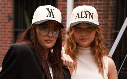 Two women wearing hats with 'NY' and 'ALYN' text, standing in front of a brick wall.